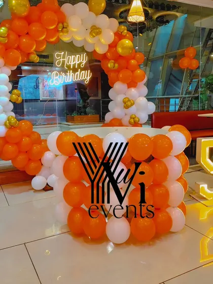 A fun orange and white balloon setup for a 5th birthday, featuring a circular arch, a balloon-covered table, and a light-up number '5'.