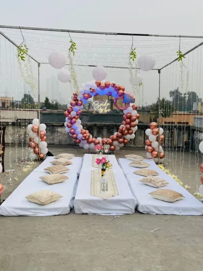 A beautiful 'Bride to be' terrace party setup. This features a metal frame with fairy lights, a circular balloon arch in rose gold and white, and a cozy low-seating arrangement, creating a perfect ambiance for a special celebration.