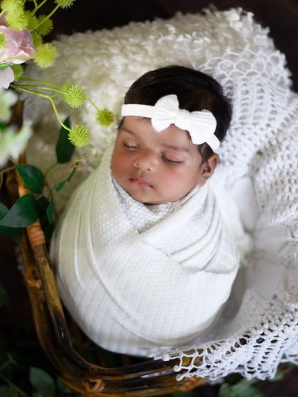 Simple and elegant. A baby girl in a white bow headband, wrapped in a textured white swaddle and placed in a rustic basket with floral accents.