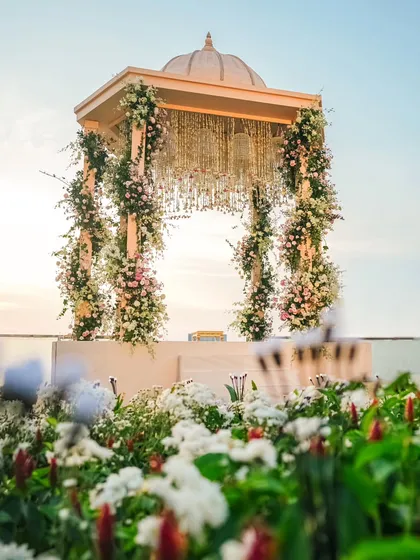 A low-angle view of the waterside Varmaala mandap, seen from behind a bed of white garden flowers. This perspective highlights the harmony between our structured design and the natural beauty of the venue.
