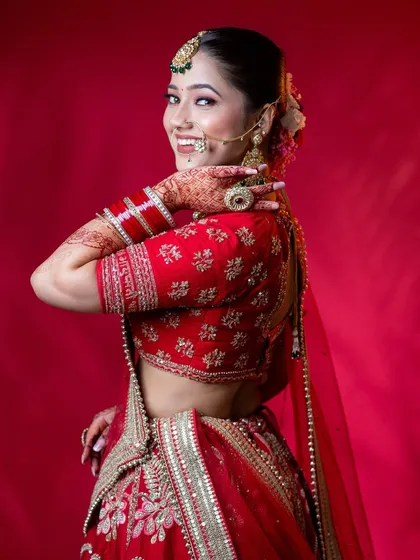 A vibrant and joyful bridal shot against a solid red background. The model's happy expression and pose showcase the complete look, including the embroidered red lehenga, traditional jewellery, and intricate henna design, perfect for demonstrating a full bridal styling.