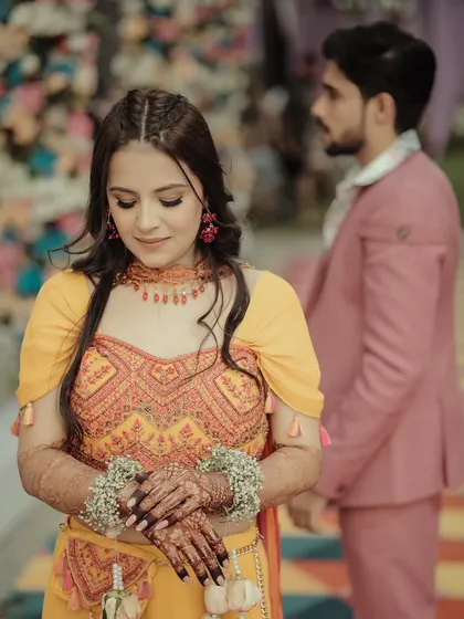 A beautiful portrait of the bride during her Haldi or Mehendi ceremony. The focus is on her serene expression and the intricate floral jewelry on her hands, with the groom softly blurred in the background.