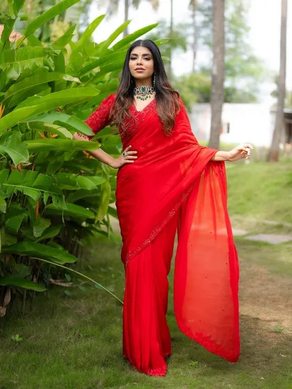 A full-length portrait of a model in a stunning red saree, set against a vibrant green garden. The color contrast makes the outfit pop, creating a visually impactful fashion shot.