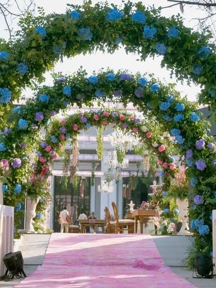 The entrance to the ceremony area, featuring a pink carpet runner leading through a series of lush floral arches in shades of blue, pink, and purple.