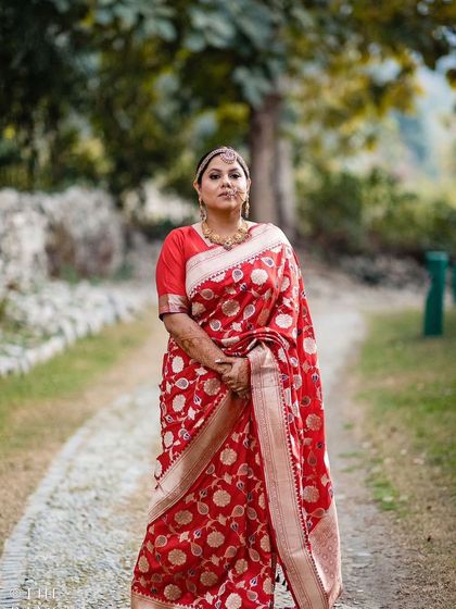 Our beautiful bride in a classic red Banarasi saree, standing in a serene outdoor setting. Her makeup is elegant and understated, allowing her natural beauty and the richness of the saree to shine.