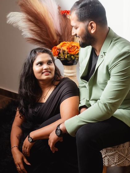 A sweet pre-wedding portrait where the couple shares a look. The warm tones of the background decor complement the green blazer and black dress perfectly.