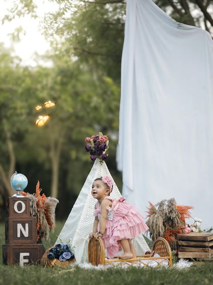 Looking up with wonder, this one-year-old enjoys her special outdoor birthday shoot. The natural setting and boho props like the teepee and dried flowers create a dreamy, adventurous vibe.