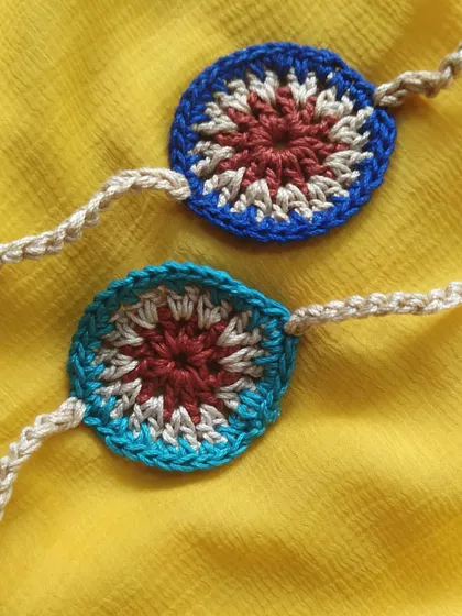 Two handmade rakhis with a rustic, 'charkha' inspired design against a bright yellow background. The intricate color work makes each piece a small work of art.