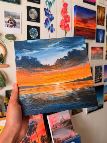 The finished orange sunset painting held in my hand, with my art wall creating a colorful and inspiring backdrop.