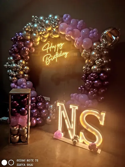 A birthday setup with light-up initials "NS" and a purple and gold balloon ring backdrop.