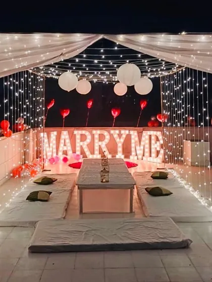 I designed this romantic rooftop proposal within a fairy light cabana. The setup includes low floor seating for an intimate dinner, heart-shaped foil balloons, and glowing 'MARRY ME' marquee letters as the centerpiece.