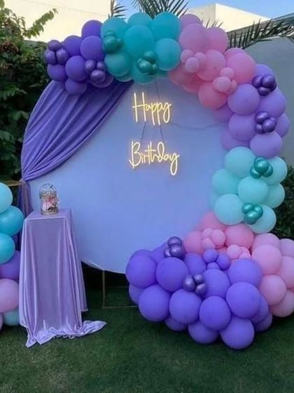 An outdoor birthday decoration featuring a circular backdrop with a neon "Happy Birthday" sign, framed by a beautiful balloon garland in purple, pink, and teal.
