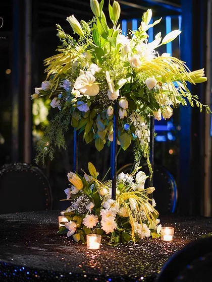 A tall, elegant centerpiece of white lilies and other blooms on a black sequined tablecloth, surrounded by candles.
