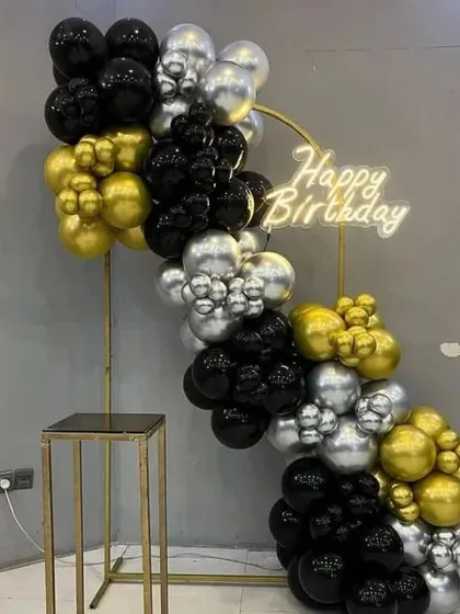 A modern and minimalist birthday setup with a black, gold, and silver balloon garland on a gold stand, complete with a neon sign.