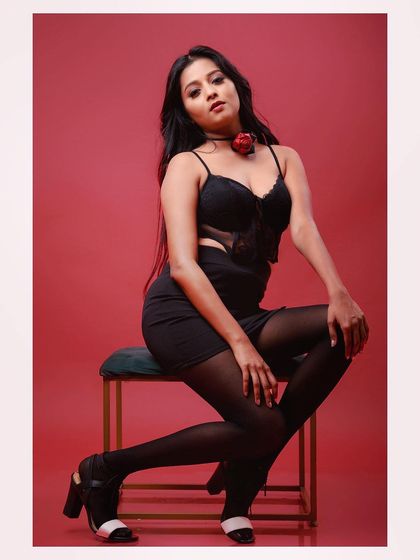 A full-length shot of a model in a glamorous black outfit against a red background. The pose is strong and confident, creating a striking fashion image.
