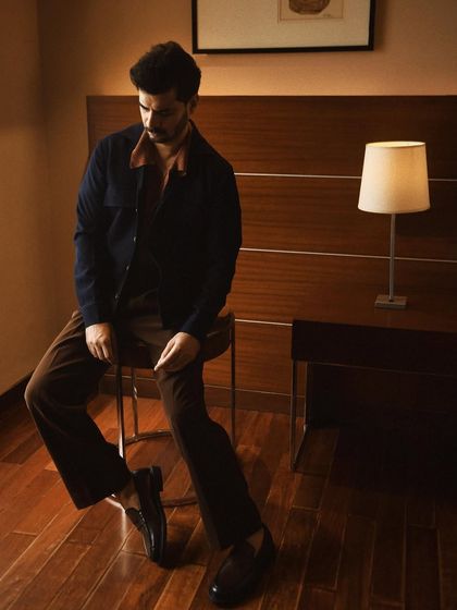 A full-length portrait of Tahir Raj Bhasin in a thoughtfully styled outfit. The composition uses the clean lines of the room to create a moody and sophisticated atmosphere.