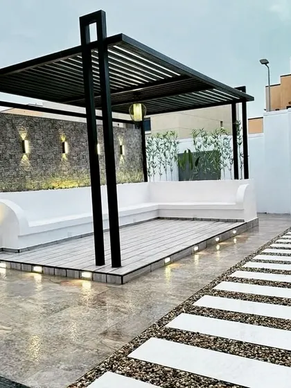 This pergola combines a sleek metal frame with a composite wood deck and a built-in white concrete bench. The back wall is finished with textured stone and accented by modern up-and-down wall lights.
