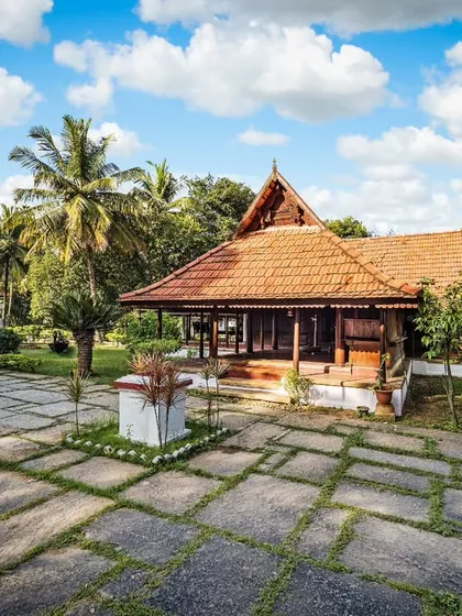 A stunning wide-angle shot of the Aranmula Palace, set against a bright blue sky. The stone-paved courtyard and surrounding palm trees complete this picture of a perfect wellness sanctuary.