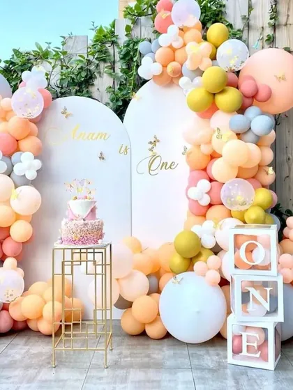 A stylish outdoor first birthday setup with a soft, warm color palette. The decoration includes dual arched backdrops, an organic balloon garland, a cake stand, and 'ONE' letter blocks.