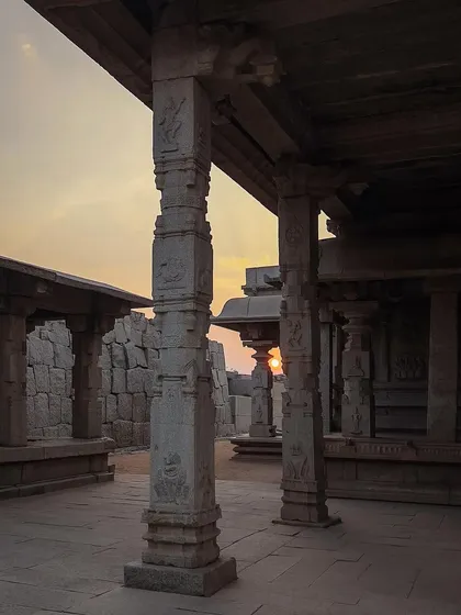 The setting sun peeks through the carved stone pillars of a temple in Hampi. The warm light and intricate details create a serene and timeless atmosphere.