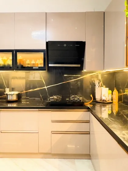 A showcase of modern luxury, this L-shaped kitchen features high-gloss beige cabinets paired with a dramatic black marble countertop and backsplash. We integrated under-cabinet lighting and glass-front display units to add depth and highlight the client's crockery.