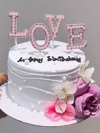 A beautiful white cake with purple flowers and a "LOVE" topper, ideal for birthdays or anniversaries.