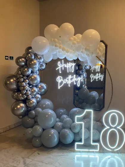 An 18th birthday setup with a grey and white balloon ring, a mirror backdrop, and neon signs.