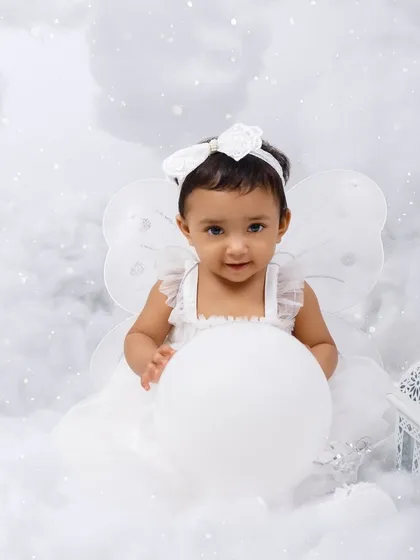A little angel sits amongst the clouds, holding a white balloon for her first birthday portrait.