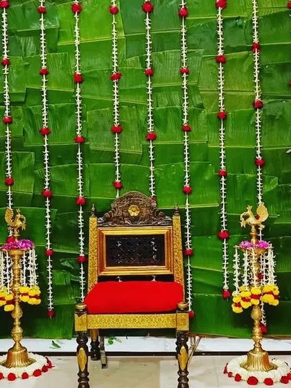 A closer look at a traditional decor setup for a home ceremony. The backdrop of fresh green leaves is adorned with strings of white and red flowers, complemented by brass lamps and baskets of marigolds.
