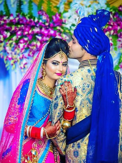 A vibrant wedding photo. The groom wears a gold and blue brocade sherwani with a royal blue stole and safa, perfectly matching the bride's colorful attire.