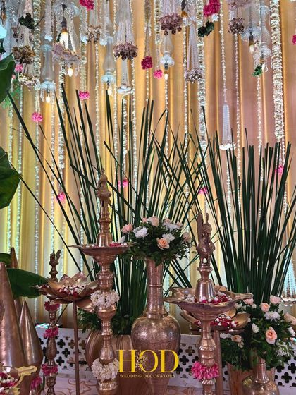 A collection of traditional brass lamps (diyas) and vessels, interspersed with fresh flowers and greenery, creating a rich and authentic decor corner.