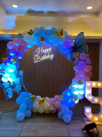 This butterfly-themed 5th birthday setup uses a balloon ring in shades of blue and pink. The large butterfly cutouts and a neon 'Happy Birthday' sign complete the magical feel.