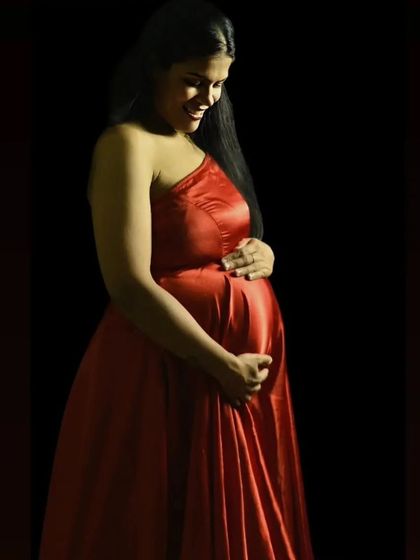 Using dramatic, low-key lighting, this portrait creates a sense of intimacy and wonder. The mother-to-be, dressed in a red gown, smiles down at her bump, beautifully illuminated against a dark background.
