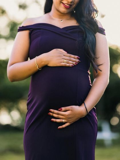 A close-up shot focusing on the mother-to-be's hands cradling her baby bump. She wears a beautiful dark purple dress, and the image exudes a sense of warmth and anticipation.