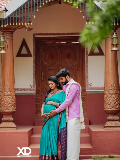 A tender moment as the couple stands together, their hands cradling the baby bump in front of their traditional home.