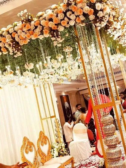 A behind-the-scenes look at a beautiful mandap being set up. The golden structure, adorned with peach and white flowers and hanging crystals, is ready for the wedding ceremony.