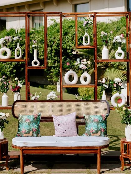 A unique and artistic photo backdrop for a modern Mehendi. This wooden shelving unit is filled with an eclectic mix of minimalist white vases and fresh flowers, offering a contemporary alternative to a traditional floral wall.