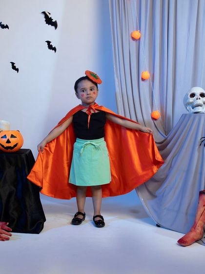 A child model in a cute pumpkin-themed Halloween costume with an orange cape and matching hair accessory.