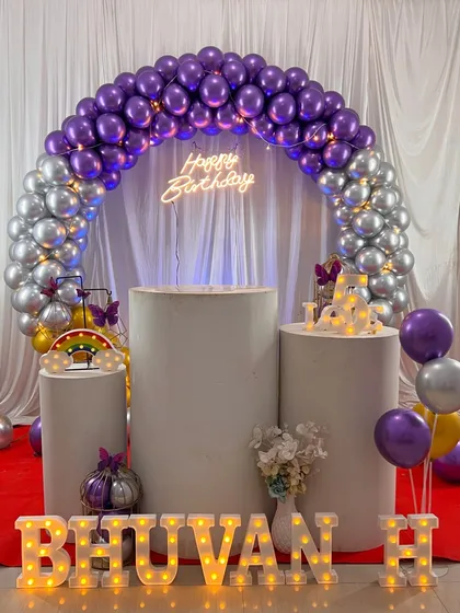 A complete birthday stage setup with a purple and silver chrome balloon ring. We included a 'Happy Birthday' neon sign, marquee letters for the name 'BHUVAN H', and pedestals for the cake.