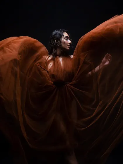 A dramatic shot with a flowing orange tulle. The model's wet-look, curly hair and glowing skin are set against a dark background, creating a high-contrast, artistic image.