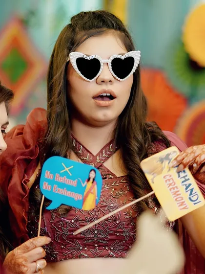 A fun and quirky shot of a guest at a Mehendi ceremony, holding props and wearing heart-shaped sunglasses.