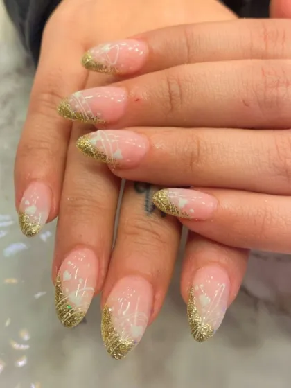 An elegant and festive design on almond-shaped nails. This look combines a sheer pink base with gold glitter tips and delicate, hand-drawn white floral details.