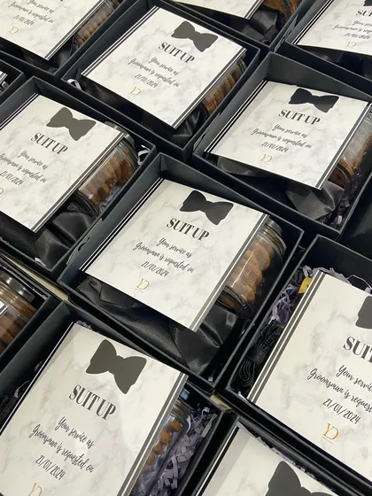 A look inside our 'Suit Up' groomsmen boxes. Each one contains a jar of treats and a formal invitation card, all set against a sleek black satin lining.