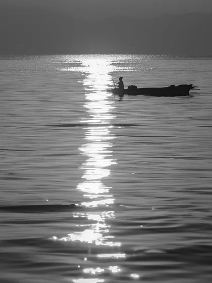 A lone fisherman in his boat is silhouetted against the glittering reflection of the sun on the water. This minimalist, high-contrast image is a peaceful study of light and water.