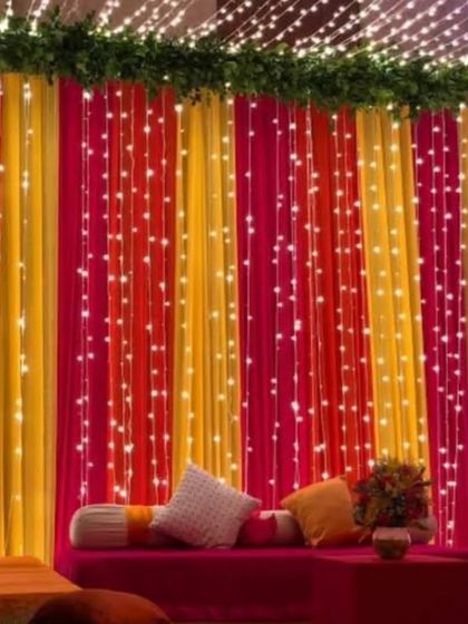 A cozy seating area with a backdrop of colorful drapes and fairy lights. The pink and yellow fabric creates a warm and inviting atmosphere for a Sangeet or Mehandi night.