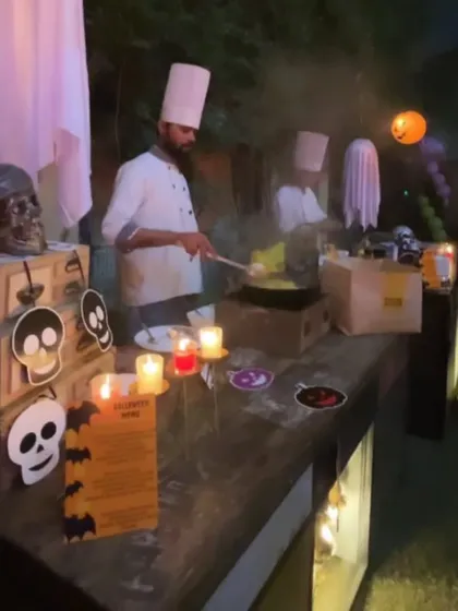 A chef creates a spooky ambiance with smoke effects at a live cooking station during a Halloween party. The station is decorated with skulls and other themed props.