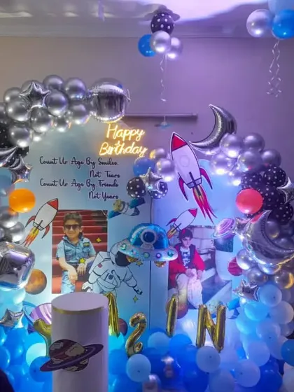 A personalized space-themed birthday party with custom photo backdrops. The setup includes a silver and blue balloon arch, rocket and moon foil balloons, and a light-up number '5'.