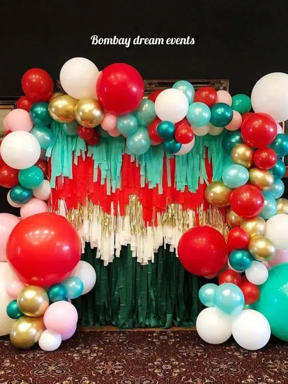 This creative Christmas backdrop uses a fringe curtain in festive colors, framed by a vibrant balloon arch with large red, green, and gold balloons for a fun and modern holiday look.