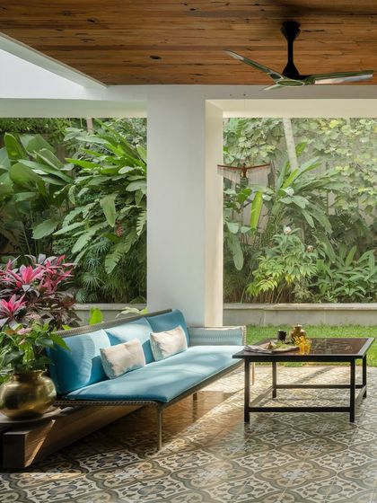 A cozy outdoor seating area surrounded by a dense tropical garden. The design uses a wooden ceiling and patterned tiles to define the space, while large openings ensure the homeowners are always connected to the green outdoors.