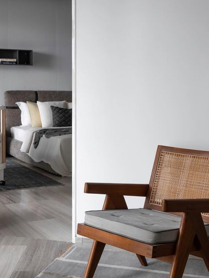A peek into a serene bedroom from the hallway. The classic Pierre Jeanneret-style chair in the foreground hints at the understated luxury and earthy, farmhouse-inspired aesthetic of the private space beyond.